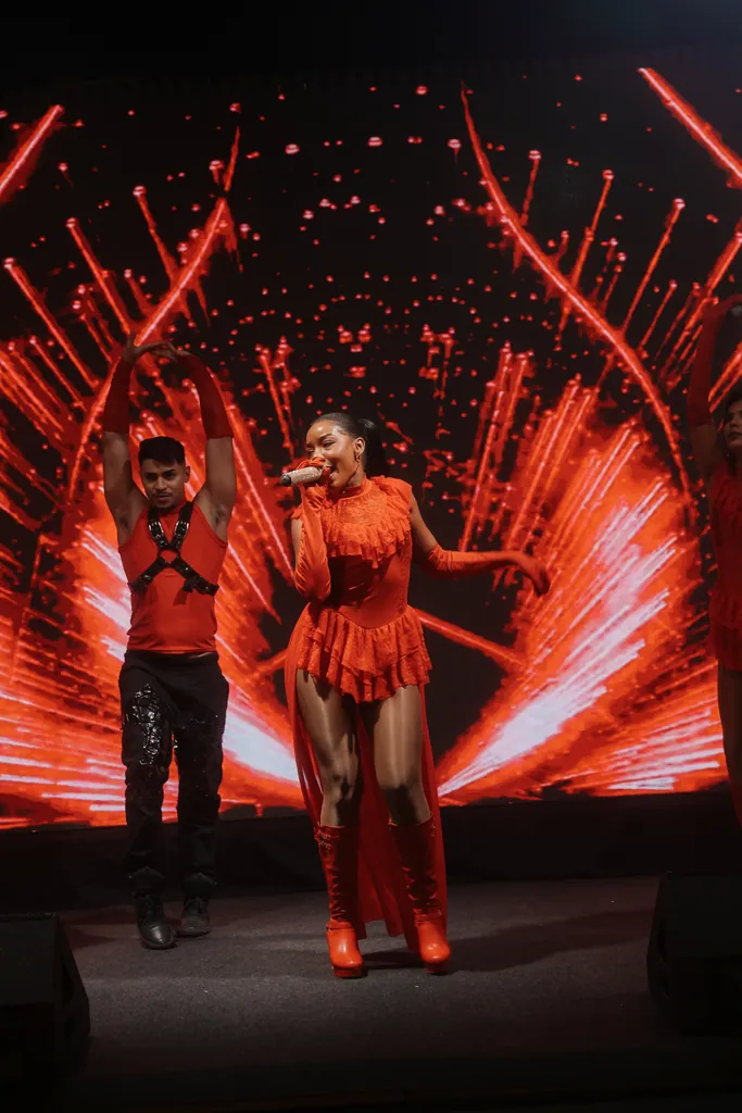 Cantora de vermelho ao microfone com artista ao lado, feixes vermelhos ao fundo